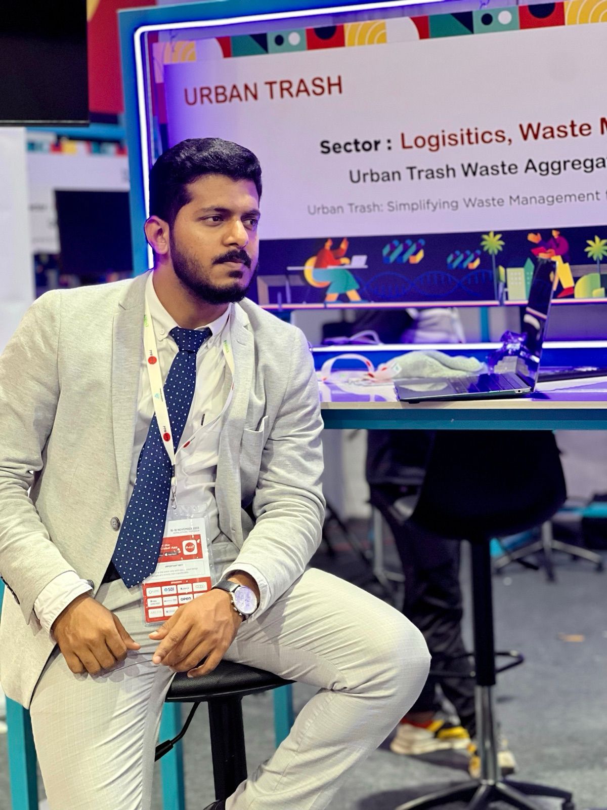 A man in a grey blazer sitting on a stool at a startup showcase stand, with an Urban Trash sign behind him.