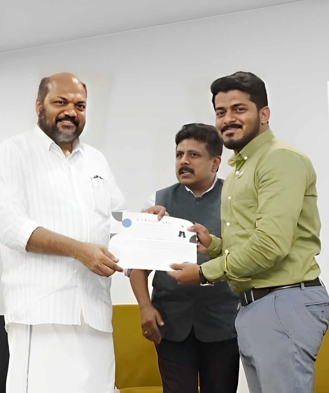 A man in a green shirt receiving a certificate from a man in white as another man in a grey waistcoat looks on.