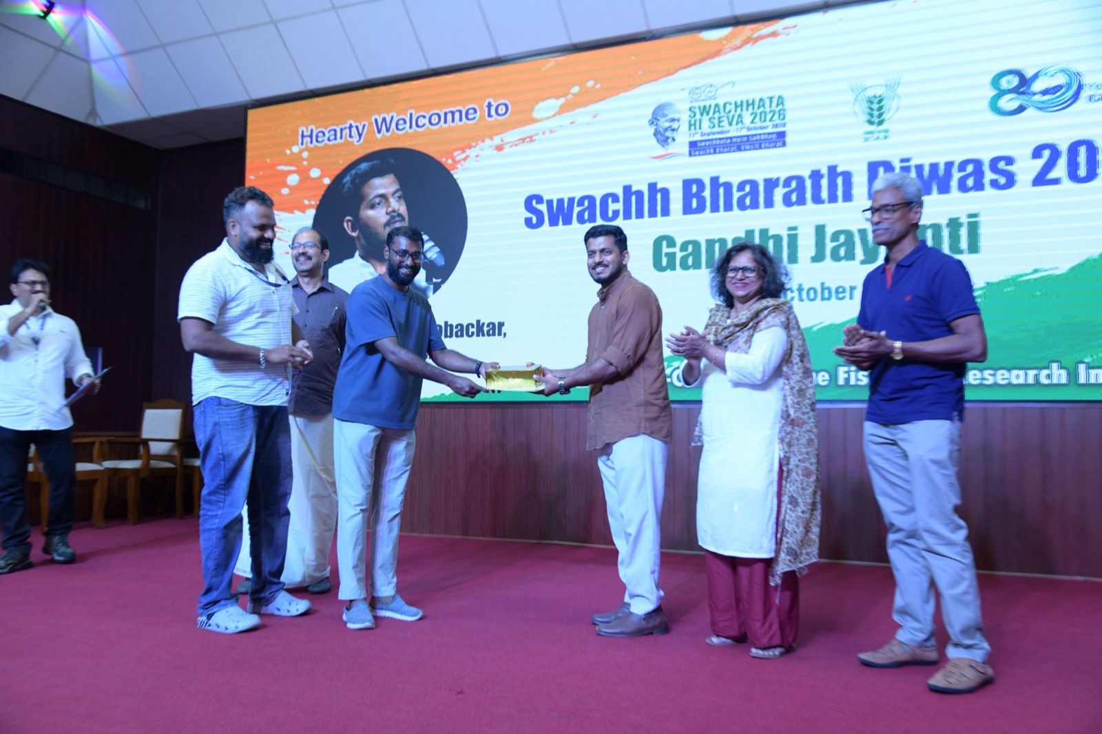 On stage, a man in a blue T-shirt hands a gold-wrapped gift box to a man in a brown shirt while guests applaud, with the Swachh Bharath Diwas 2026 screen behind them.