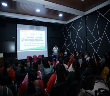A speaker presenting to a seated audience in a hall, with a slide on the screen.