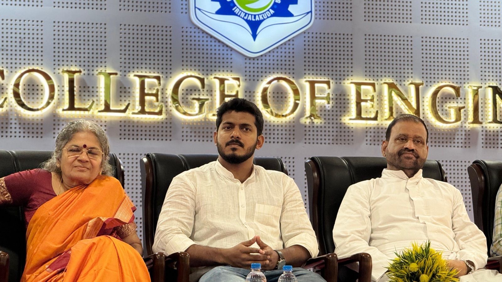A man in a white shirt seated between two guests on a stage, under a lit sign for a college of engineering.
