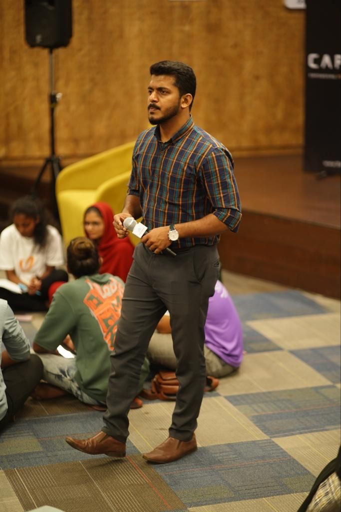 A man in a checked shirt standing with a microphone among people sitting on the floor at a founders workshop.