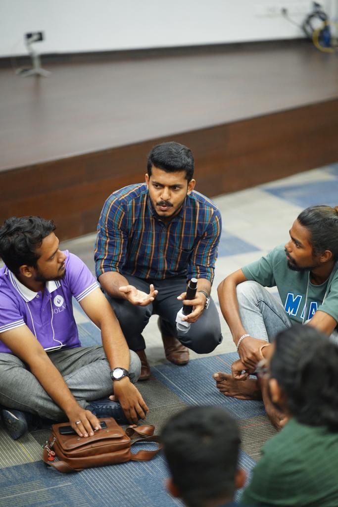 A man in a checked shirt crouching to talk with a microphone to a circle of founders seated on the floor.