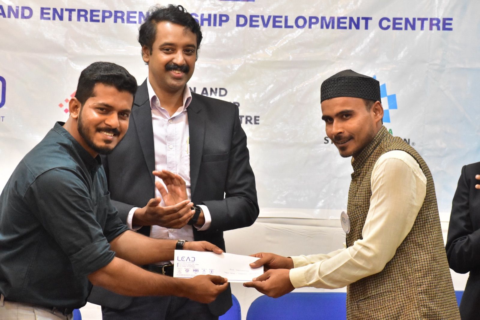 Two men smiling and holding a paper between them while a third man in a suit applauds, in front of an IEDC banner.