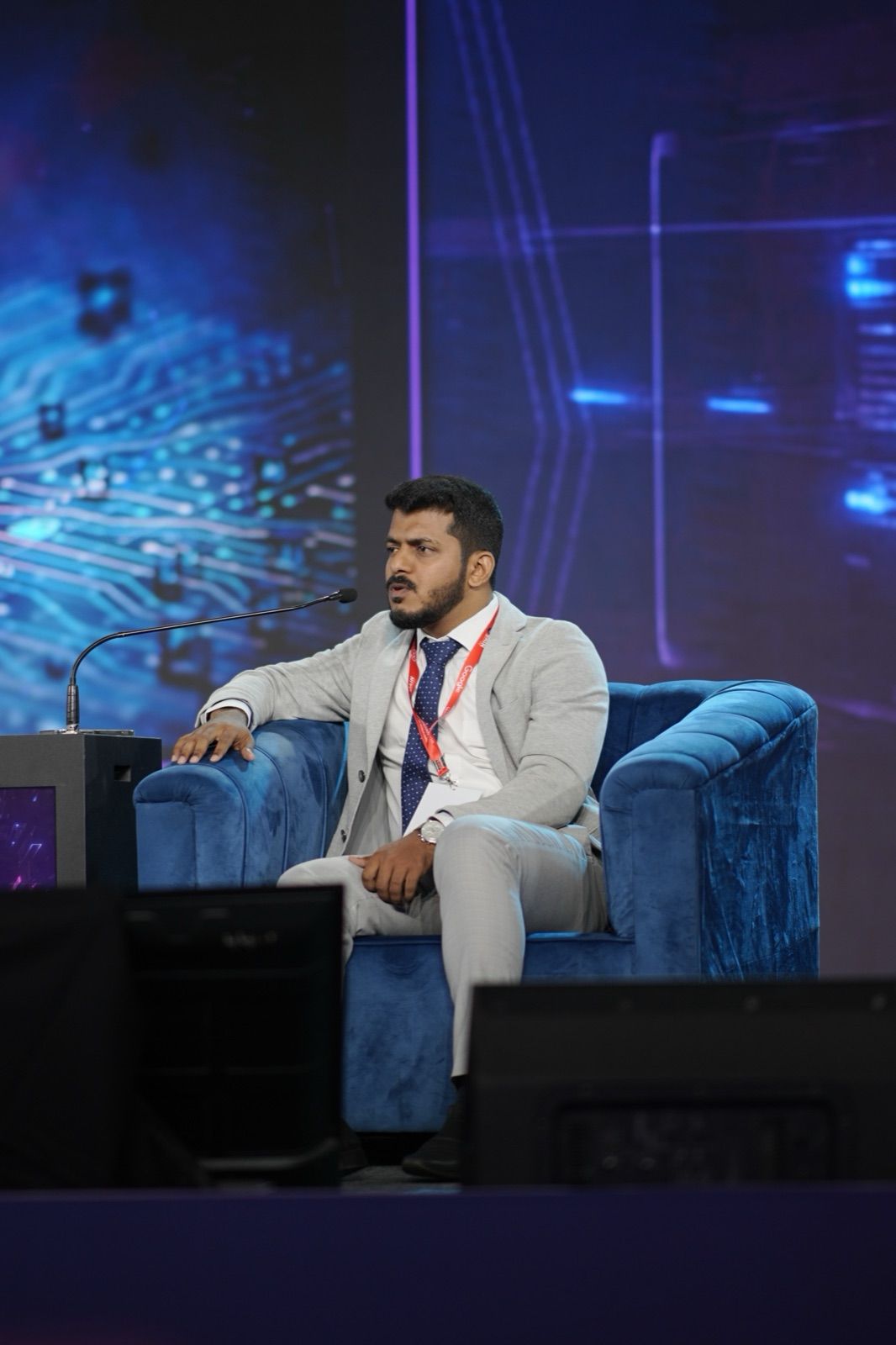 A man in a grey suit seated on a blue sofa on a stage, with a lit circuit-board backdrop.