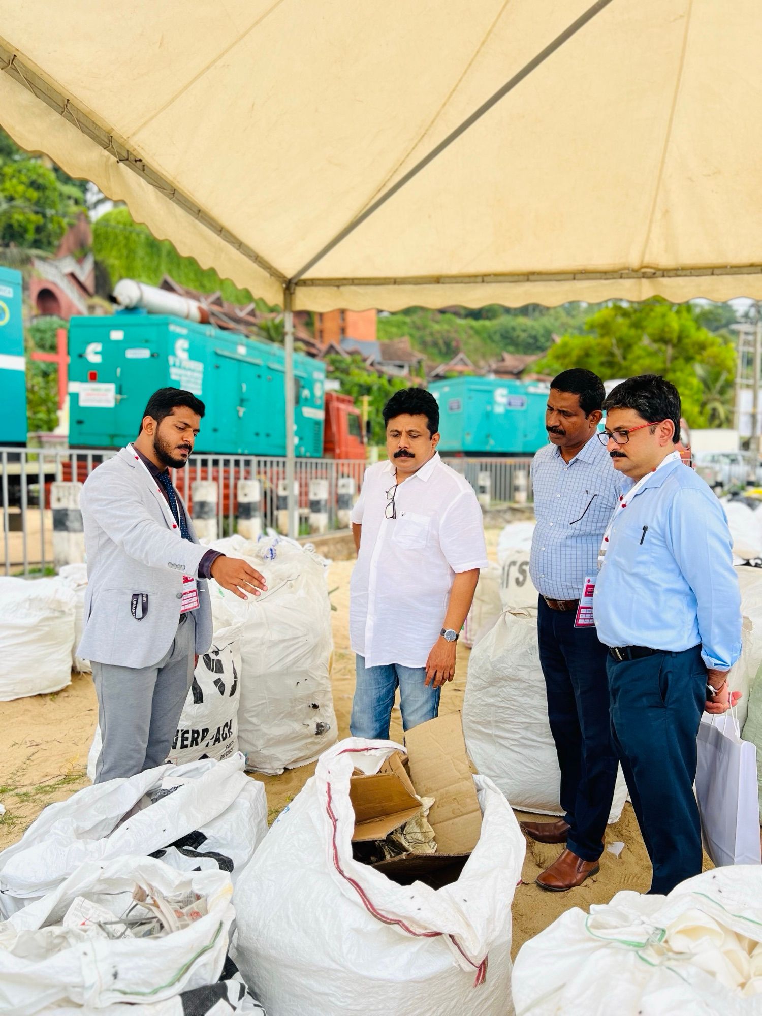 A man in a light grey jacket points to large white sacks of sorted waste under a tent while three men look on.
