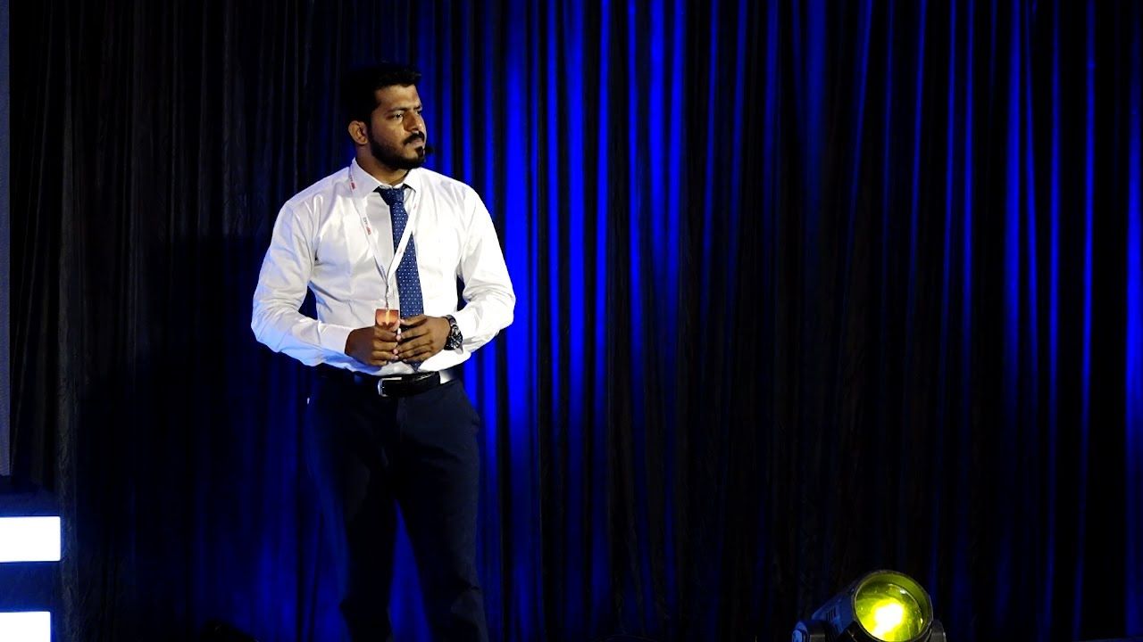 A speaker in a white shirt and dark tie standing on a stage with a deep blue curtain behind him.