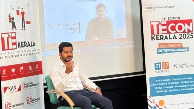 A speaker seated on a stage chair, mid-sentence, between event banners, with a projector screen behind him.